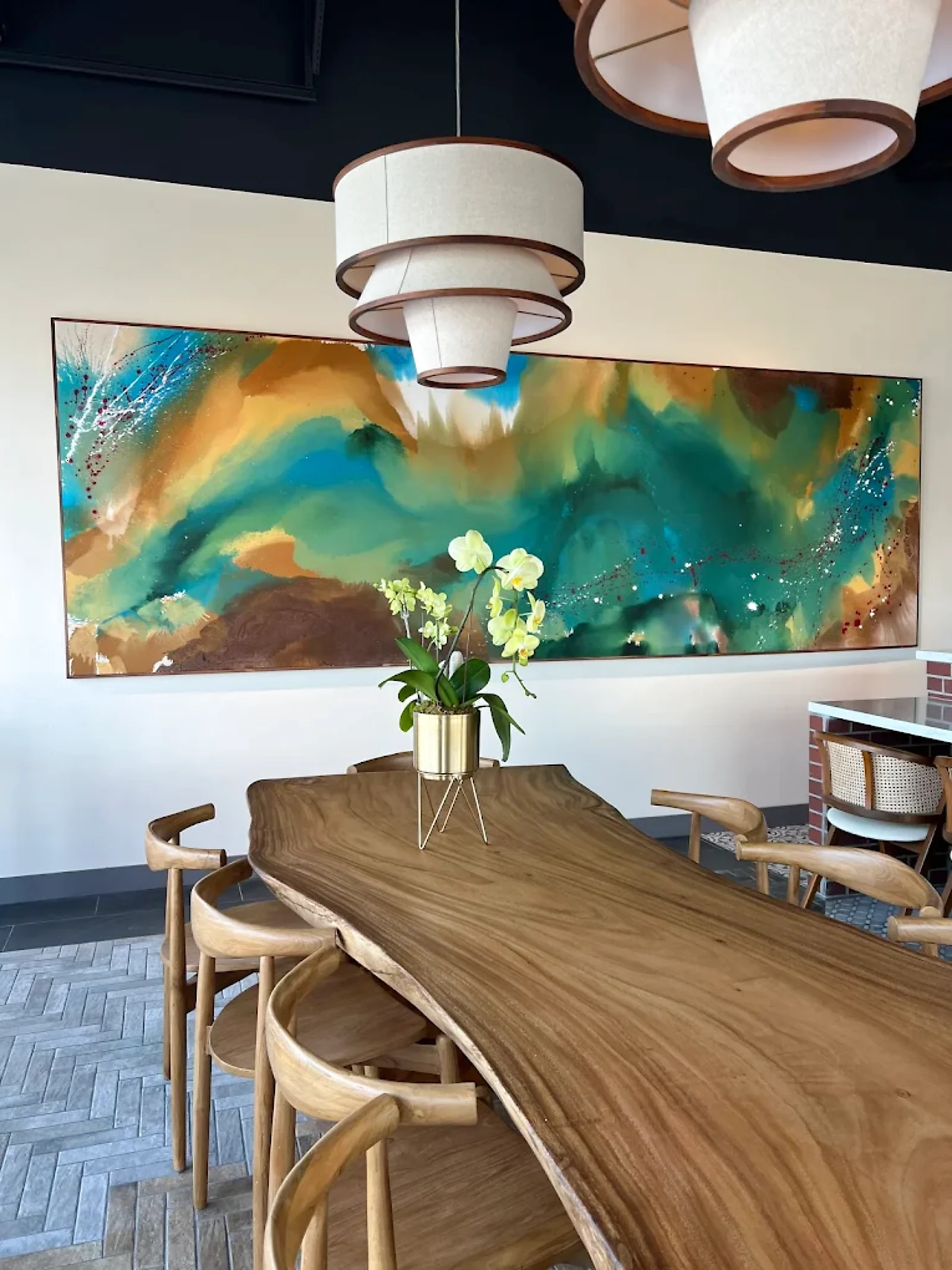 The long live-edge communal table at Between Us in warm morning light, beneath a wall-spanning teal and copper painting